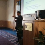 A man gestures next to a podium and in front of a screen